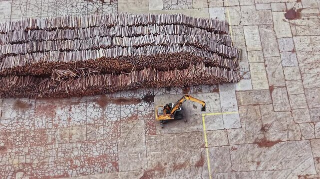 An excavator handles tree branches beside rows of stacked timber on a paved work site, seen from above during the day.