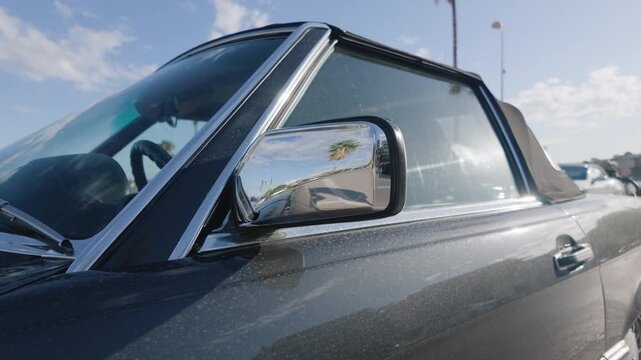 Dark classic convertible with a black soft top and a chrome side mirror reflecting a palm tree on a sunny day, French Riviera, France.