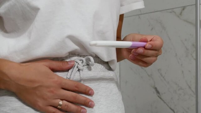 Close-up of woman holding pregnancy test and touching lower abdomen in bathroom, pregnancy and reproductive health concept