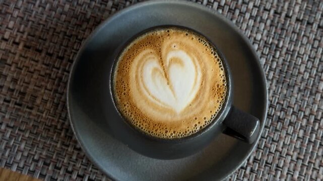 Overhead view of a black coffee cup with heart shaped latte art on a matching saucer, resting on a woven placemat and wooden table