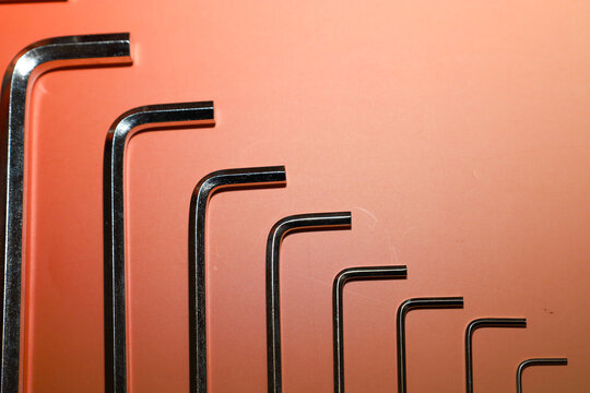 Set of metal hex keys arranged by size on a bright orange and peach gradient background. Chromed tools are laid out in a clear vertical row, creating a concise graphic composition.