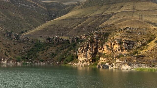 Calm lake water moves gently below rugged illuminated cliffs and rolling brown hills, creating a peaceful mountain landscape with natural reflections