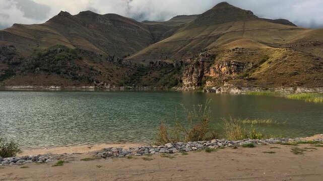 Calm lake water moves gently below rugged illuminated cliffs and rolling brown hills, creating a peaceful mountain landscape with natural reflections