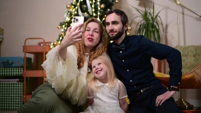 A mother, father and their young daughter sit together on the floor beside a decorated Christmas tree and take a selfie with a smartphone. 