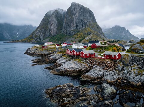 Nordic fishing village with red wooden cabins on a rocky coast. Bright red stilt houses with white trim line a rugged shoreline of layered gray boulders, while a steep granite mountain rises behind