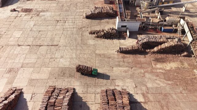 A green forklift crosses a paved timber yard between rows of stacked lumber beside work buildings, viewed from above during daylight.