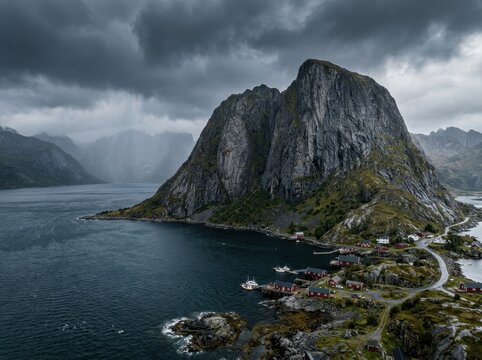 Rugged rocky mountain beside a fjord under stormy clouds, landscape photography. A steep gray granite peak rises over a small coastal village with red cabins, distant mountains fade in the haze, and