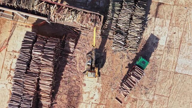 An excavator moves timber beside rows of stacked logs at a wood processing yard, seen from above during the day.