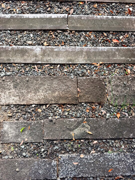 Overhead view of weathered rectangular stone slabs arranged in parallel rows, separated by gravel and scattered fallen leaves, highlighting rough textures, cracks, and earthy tones.

