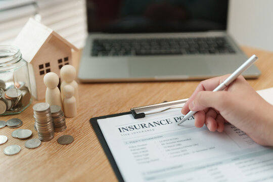 Hand signing on insurance policy form clipboard beside a wooden house, family figures and stacked coins, connecting property coverage, family protection and financial planning at a desk.