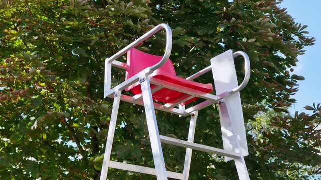 Empty aluminum umpire chair with pink seat stands under a leafy tree in sunlight.