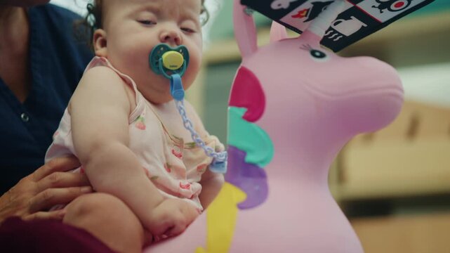 Low angle view of a baby girl with a pacifier sitting on a pink unicorn hopper with a black and white sensory book on its head, French Riviera, France.