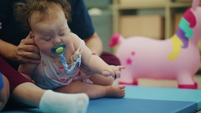 Baby girl with a pacifier sitting on a blue mat between a therapist's legs, a pink unicorn hopper in the background, French Riviera, France.