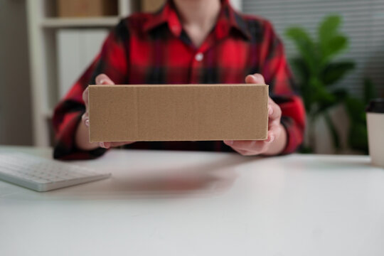 Small business owner holding a cardboard parcel while working at a computer, managing online orders and preparing products for shipment from a home office.