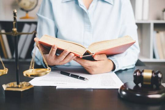 Person reading an open reference book at a desk with scales of justice, a wooden gavel, and printed documents, illustrating legal research, case preparation, office study,