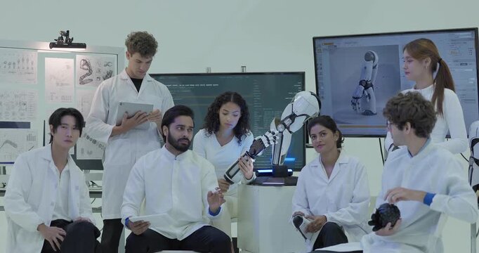 Engineers in white lab coats sit in a row watching a large screen in front of them, one holding up the robotic arm as colleagues with the gripper and test balls discuss the design.