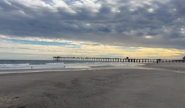 Down tilt view from dramatic cloudy sky and pier over ocean to a sandy beach with copy space
