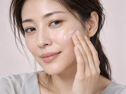 スキンケアで頬に白いクリームを優しく塗り広げる上品な女性 透明感のある艶肌と自然な微笑みが映える淡いピンク背景の美容クローズアップ