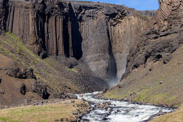 Tall basalt columns frame Litlanesfoss waterfall in East Iceland, with spray rising from the narrow gorge and whitewater flowing over rocks between green summer slopes. © Artem