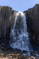Water cascades down Studlafoss waterfall between towering basalt columns in East Iceland. Sunlit spray, scattered boulders and a clear blue sky frame this summer landscape. © Artem