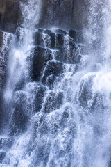 White cascades and fine spray break across dark basalt columns at Studlafoss in East Iceland, creating a layered water texture that fills the vertical frame as a natural background. © Artem