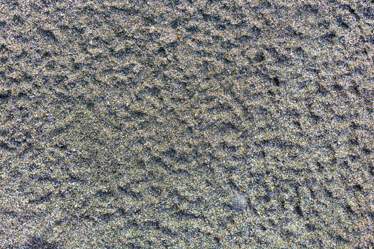 Close up image of textured dark river sand displaying fine granular particles and subtle wind ripples, compact dark sandy ground showing subtle micro ridges and coarse mineral grains