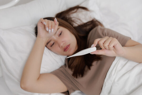 Sick Asian woman lying in bed feeling feverish checking thermometer with tired and worried expression in bedroom