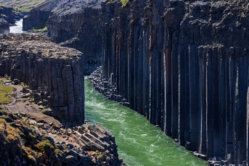 Green water flows between tall basalt columns in Studlagil canyon, East Iceland. The Jokla river curves past rocky banks and sparse summer vegetation in Jokuldalur. © Artem