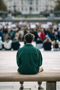 Young adult sitting on a wooden bench facing a blurred crowd in a public square outdoors