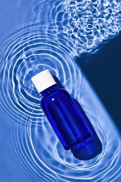 Transparent cobalt blue glass bottle with white cap resting in clear water, surrounded by concentric ripples and bright reflections in a top view aquatic scene