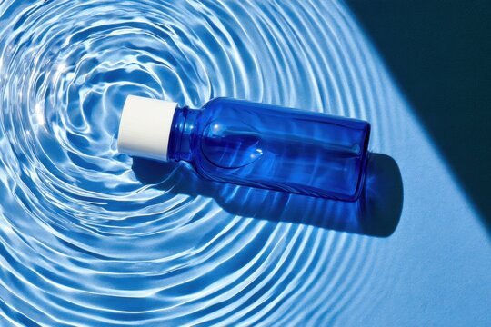 Transparent cobalt blue glass bottle with white cap floating on clear water amid concentric ripples, overhead composition with vivid blue tones and shadow