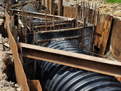Black corrugated drainage pipe in excavated trench with shoring supports and surrounding soil