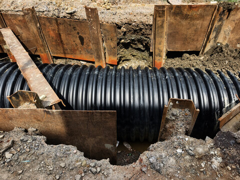 Black corrugated drainage pipe in excavated trench with shoring supports and surrounding soil