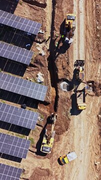 An aerial view shows excavators and dump trucks moving soil beside rows of solar panels at a solar farm during daylight.