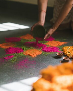 Close-up of a woman's hands pouring vivid pink, orange and green powder to make a rangoli beside marigolds and clay lamps