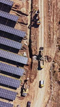 Workers guide earthmoving machines along a dirt track beside rows of solar panels at a solar farm construction site, seen from above during daytime.