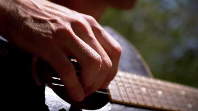 Guitarist fingerpicks acoustic guitar strings during a sunny outdoor session in selective focus. Warm sunlight highlights hand movement, evoking peaceful creative expression and leisure.