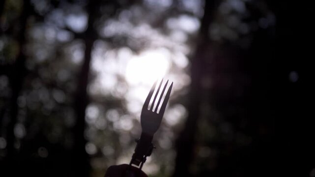 Hand holds a silhouette of a metal camping fork backlit by bright sun through forest canopy. High contrast lighting creates strong outlines ideal for outdoor gear promotions.