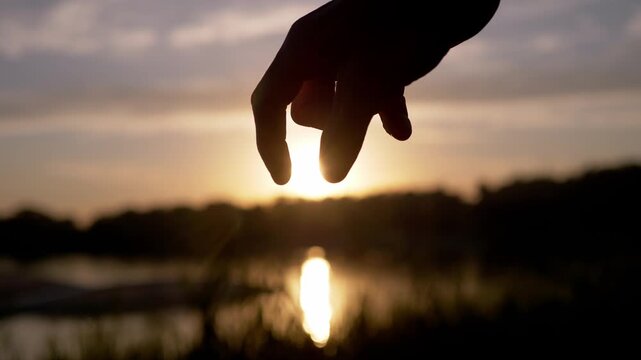 Silhouette hand pinching setting sun over lake shoreline in close-up. Tight framing emphasizes fingertips holding the golden sunset light above calm water reflections.