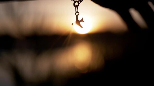 An airplane pendant silhouette dangles against a warm golden hour sunset. Radiant sun flare and deep bokeh create high contrast across the tranquil evening lake horizon.
