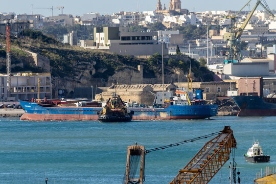 Valletta, Malta - October 14th, 2026 Cargo Ship and Tugboat Docked in Industrial Port Harbor With City Hillside Background