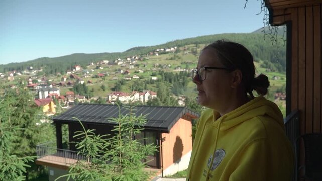 A young woman yellow hoodie glasses smokes electronic cigarette, exhaling vapor while enjoying view mountains forest warm, cloudless morning. numerous private vacation homes visible mountainside