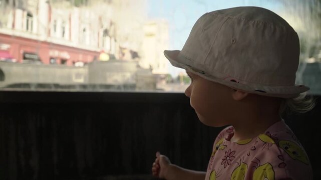 A little girl in pink clothes and a bucket hat looks around, and points her finger inside a fountain ,where water is falling in a straight stream.