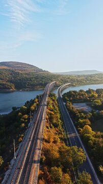 Railway tracks and a highway run side by side through trees beside a lake, with wooded hills in the distance during autumn daylight.
