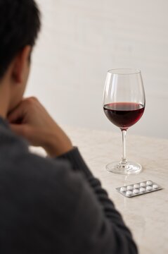 Glass of red wine beside a blister pack of tablets, viewed by a seated person. Antidepressants and alcohol use disorder. Medication interaction and mental health treatment.