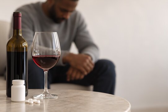 Wine bottle, glass and pills beside seated person. Antidepressants and alcoholism concept. Alcohol use disorder and mental health treatment. Medication safety and substance abuse awareness.
