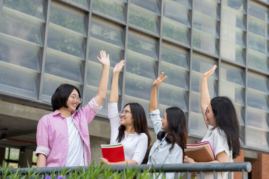 Four female university students share a high five outside a modern campus building, expressing friendship, teamwork, unity, confidence, student life, and academic achievement.