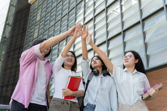 Asian university students celebrate with a group high five outdoors on campus, with books, headphones, and a laptop supporting themes of friendship, teamwork, education, and student lifestyle.