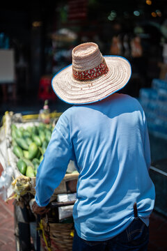 Vendor selling tropical fruits from cart on streets in China town in Bangkok, Thailand. Unrecognizable man in straw hat sells exotic mango from pushcart for customers while walking through city.