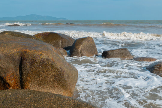 Large weathered coastal boulders surrounded by foamy ocean waves with distant mountains under clear blue sky